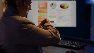 Mulher no escritório à noite a olhar para o relógio inteligente no pulso, em frente a um monitor com gráficos.
